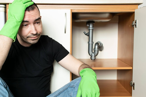 Homeowner wearing latex gloves sitting next to open cabinet under the sink worried about plumbing emergency.